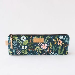 Peut inclure: Une trousse rectangulaire avec un motif floral dans les tons de bleu, vert et rose. La trousse a une fermeture éclair noire et une petite étiquette en cuir. L'étiquette sur la trousse indique "PINE STREET".