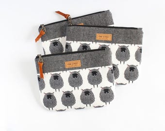 May include: Three rectangular zippered pouches featuring a white fabric base with a repeating pattern of black sheep. Each pouch has a dark gray felt-like top section, a black zipper with a brown leather pull, and a tan rectangular label reading PINE STREET.