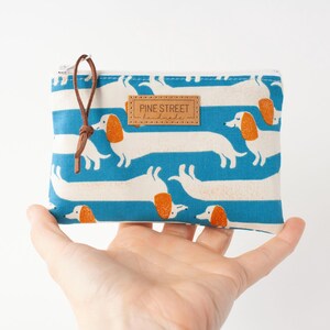 May include: A rectangular, zipped pouch with a blue and white striped pattern featuring white dachshund dogs with orange ears. The pouch has a brown leather pull and a small leather tag that reads "PINE STREET handmade."