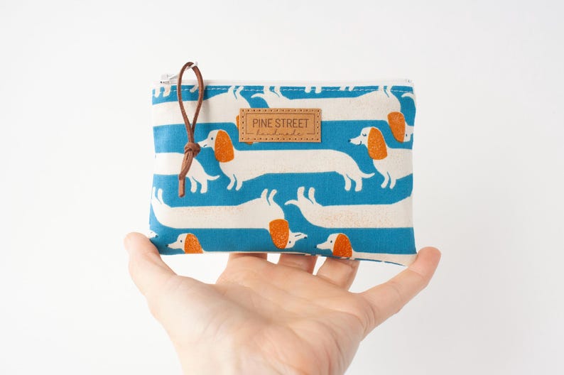 May include: A rectangular, zipped pouch with a blue and white striped pattern featuring white dachshund dogs with orange ears. The pouch has a brown leather pull and a small leather tag that reads "PINE STREET handmade."