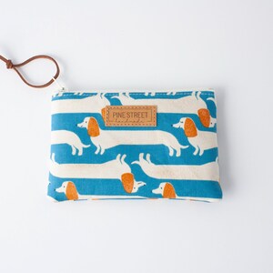 May include: A rectangular coin purse with a blue and white striped pattern featuring dachshund illustrations. The purse has a brown leather tag that reads "PINE STREET handmade" and a brown leather loop attached to the zipper.