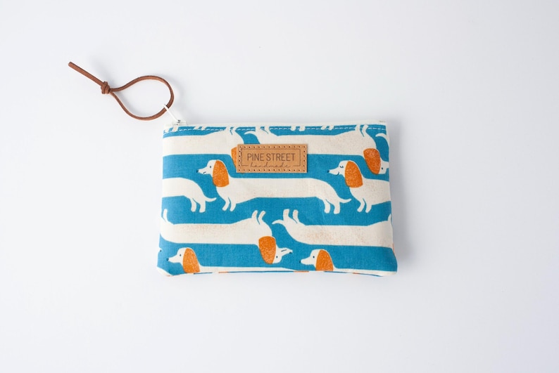 May include: A rectangular coin purse with a blue and white striped pattern featuring dachshund illustrations. The purse has a brown leather tag that reads "PINE STREET handmade" and a brown leather loop attached to the zipper.