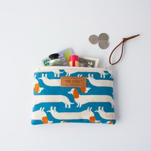 May include: A zipped pouch featuring a blue pattern of white dachshunds with orange ears. The pouch is open, revealing contents like a gift card, lip balm, and coins. The label reads "PINE STREET".