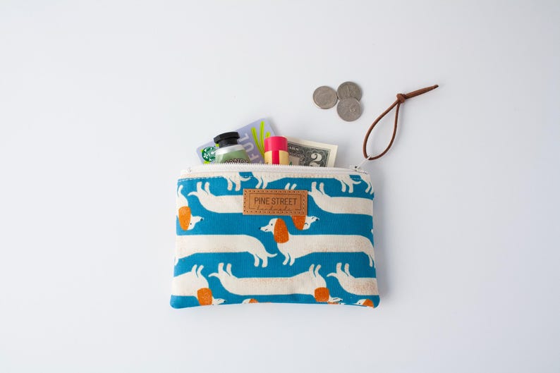 May include: A zipped pouch featuring a blue pattern of white dachshunds with orange ears. The pouch is open, revealing contents like a gift card, lip balm, and coins. The label reads "PINE STREET".