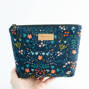 Peut inclure: Une pochette zipp&eacute;e bleu marine fonc&eacute; avec un imprim&eacute; botanique color&eacute; de petites fleurs orange, roses et bleu clair sur un feuillage vert. Une &eacute;tiquette rectangulaire en cuir beige avec le texte PINE STREET handmade est centr&eacute;e sur le devant.