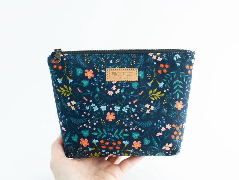 Peut inclure: Une pochette zipp&eacute;e bleu marine fonc&eacute; avec un imprim&eacute; botanique color&eacute; de petites fleurs orange, roses et bleu clair sur un feuillage vert. Une &eacute;tiquette rectangulaire en cuir beige avec le texte PINE STREET handmade est centr&eacute;e sur le devant.