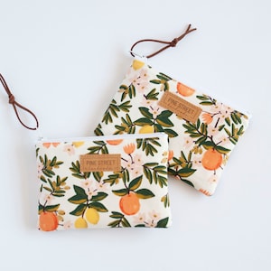 May include: Two small zippered pouches with a white background and a floral print featuring oranges, lemons, and green leaves. The pouches have a brown leather pull tab and a brown leather label that reads "PINE STREET" in white text.