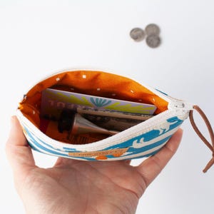 May include: A small, open zippered pouch with a blue and white striped pattern, held in a hand. The interior is orange with white polka dots. The pouch contains a tube of cream, cards, and cash. A brown leather pull is attached to the zipper.