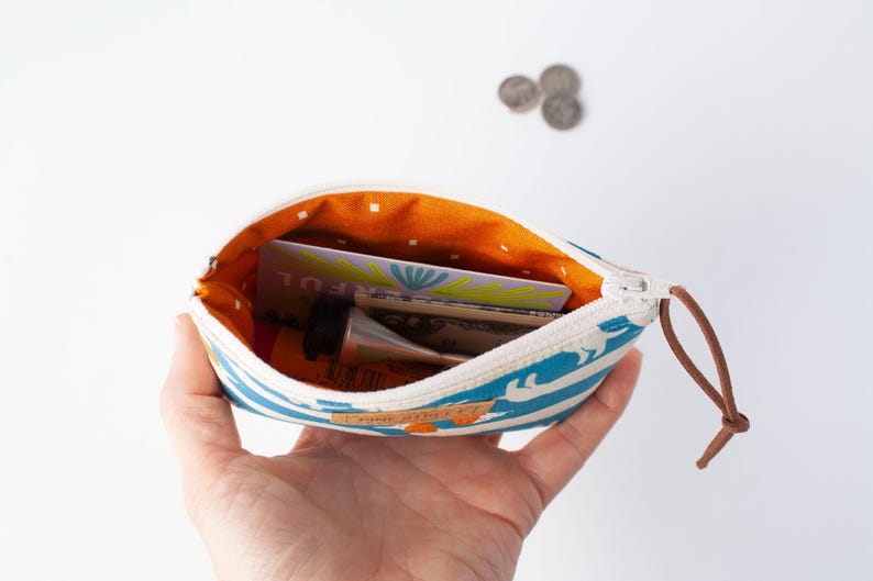 May include: A small, open zippered pouch with a blue and white striped pattern, held in a hand. The interior is orange with white polka dots. The pouch contains a tube of cream, cards, and cash. A brown leather pull is attached to the zipper.