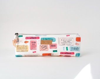 May include: A rectangular white pencil case with a zipper closure and a leather brand tag labeled Pine Street. The surface features a colorful print of various rectangular shapes resembling candy wrappers with text including Awesome, Super, Smart, and You Are No. 1.