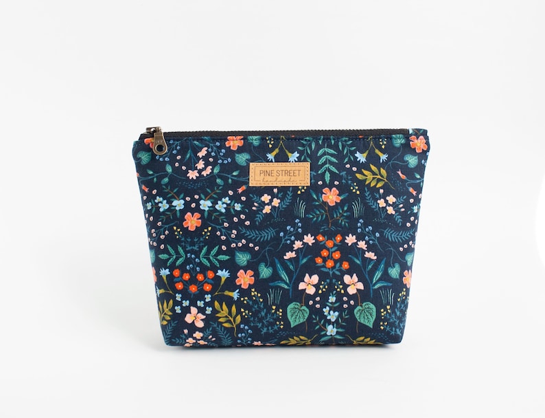 Peut inclure: Une pochette zipp&eacute;e bleu marine fonc&eacute; avec un motif r&eacute;p&eacute;titif de fleurs, de feuilles et de vignes color&eacute;es dans des tons orange, bleu clair, rose et vert. Une &eacute;tiquette rectangulaire beige avec le texte PINE STREET handmade est centr&eacute;e.