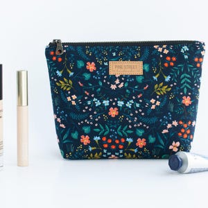 Peut inclure: Une pochette en tissu bleu fonc&eacute; avec un motif floral color&eacute;, une fermeture &eacute;clair noire et une &eacute;tiquette en cuir beige portant l'inscription Pine Street Handmade. &Agrave; c&ocirc;t&eacute; se trouvent un fond de teint bareMinerals, un tube dor&eacute; et un tube en m&eacute;tal.