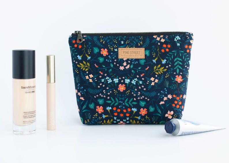 Peut inclure: Une pochette en tissu bleu fonc&eacute; avec un motif floral color&eacute;, une fermeture &eacute;clair noire et une &eacute;tiquette en cuir beige portant l'inscription Pine Street Handmade. &Agrave; c&ocirc;t&eacute; se trouvent un fond de teint bareMinerals, un tube dor&eacute; et un tube en m&eacute;tal.