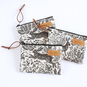 May include: Four small zippered pouches with a black and white rabbit and floral print. Each pouch has a brown leather tag that reads "PINE STREET" and a brown leather loop attached to the zipper.