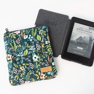 May include: A dark green quilted fabric sleeve featuring a colorful floral and botanical print with a zipper closure and a small tan rectangular label reading Pine Street. Beside it is a black e-reader in a dark gray textured case displaying a book cover.