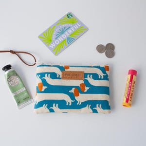 May include: A flat lay featuring a cream-coloured pouch with a dachshund pattern, a tube of hand cream, a lip balm, a gift card, and coins. The pouch has a brown leather tag that says "PINE STREET". The gift card says "WONDERFUL".