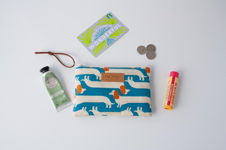 May include: A flat lay featuring a cream-coloured pouch with a dachshund pattern, a tube of hand cream, a lip balm, a gift card, and coins. The pouch has a brown leather tag that says "PINE STREET". The gift card says "WONDERFUL".