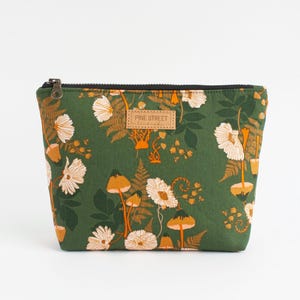 Peut inclure: Une pochette zippée vert forêt avec un motif répétitif de fleurs blanches, de champignons orange et de feuilles de fougère. Une étiquette rectangulaire beige avec le texte PINE STREET handmade est cousue au centre du panneau avant.