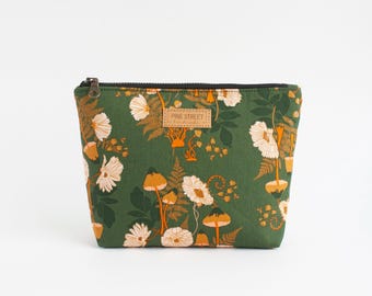 Könnte beinhalten: Eine waldgrüne Reißverschlusstasche mit einem sich wiederholenden Muster aus weißen Blumen, orangefarbenen Pilzen und Farnblättern. Ein rechteckiges beiges Etikett mit dem Text PINE STREET handmade ist mittig auf der Vorderseite aufgenäht.
