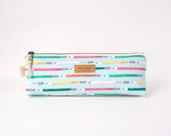 May include: A rectangular pencil case with a light blue background featuring a repeating pattern of colorful pencils in pink, yellow, and green. A brown rectangular label with the text Pine Street Handmade is centered on the front.