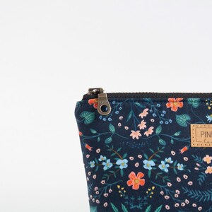 Peut inclure: Une pochette zipp&eacute;e bleu marine fonc&eacute; orn&eacute;e d'un motif floral color&eacute; avec des fleurs bleu clair, roses et orange ainsi que du feuillage vert. Une &eacute;tiquette rectangulaire en similicuir beige est cousue avec le texte PINE STREET handmade.
