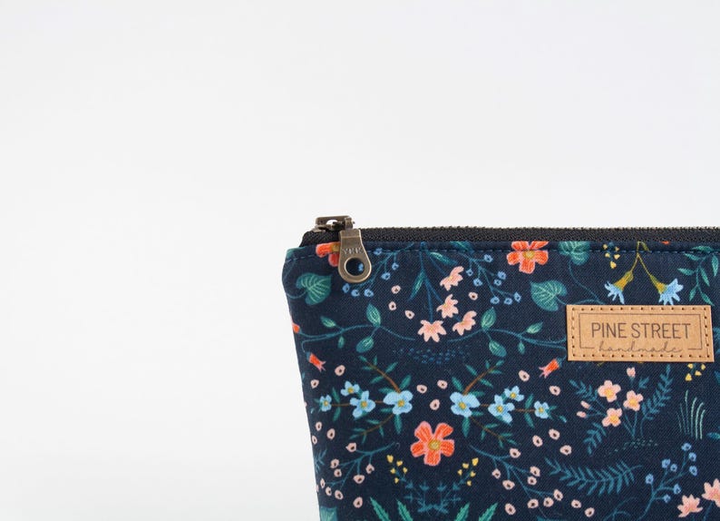 Peut inclure: Une pochette zipp&eacute;e bleu marine fonc&eacute; orn&eacute;e d'un motif floral color&eacute; avec des fleurs bleu clair, roses et orange ainsi que du feuillage vert. Une &eacute;tiquette rectangulaire en similicuir beige est cousue avec le texte PINE STREET handmade.
