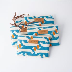 May include: Several zippered pouches featuring a repeating pattern of white dachshund dogs with orange ears and blue stripes. Each pouch has a brown leather zip pull and a small label that reads "PINE STREET handmade".