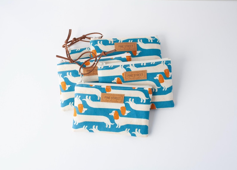 May include: Several zippered pouches featuring a repeating pattern of white dachshund dogs with orange ears and blue stripes. Each pouch has a brown leather zip pull and a small label that reads "PINE STREET handmade".