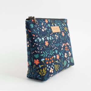 Peut inclure: Une pochette rectangulaire &agrave; fermeture &eacute;clair avec une base en tissu bleu marine fonc&eacute; orn&eacute;e d'un motif r&eacute;p&eacute;titif de petites fleurs, de feuilles et de vignes dans des tons orange, bleu clair, rose et vert. Une &eacute;tiquette beige indique Pine Street.