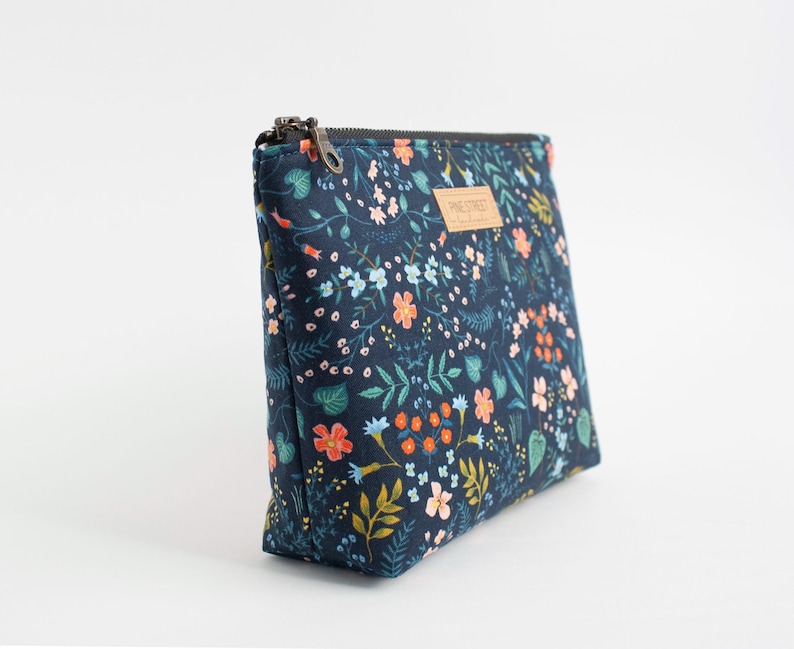 Peut inclure: Une pochette rectangulaire &agrave; fermeture &eacute;clair avec une base en tissu bleu marine fonc&eacute; orn&eacute;e d'un motif r&eacute;p&eacute;titif de petites fleurs, de feuilles et de vignes dans des tons orange, bleu clair, rose et vert. Une &eacute;tiquette beige indique Pine Street.