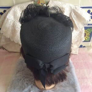 May include: A black straw hat with a black ribbon tied in a bow at the back. The hat has a black lace trim around the crown.