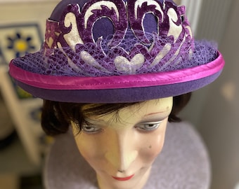 Puede incluir: Un sombrero de fieltro morado con un diseño bordado blanco y morado y una cinta rosa alrededor del borde. El sombrero está decorado con un velo morado transparente.