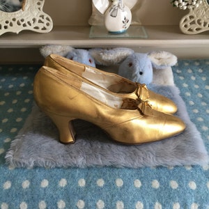 May include: A pair of gold high-heeled shoes with a bow detail on the front. The shoes are sitting on a blue and white polka dot blanket.