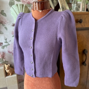 May include: A purple knitted cardigan with a button-up front and puffed sleeves. The cardigan is on a mannequin with a brown skirt.