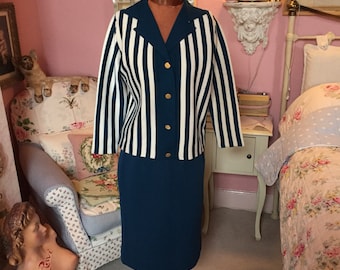 Puede incluir: Una chaqueta vintage a rayas azul y blanca con botones dorados y una falda azul verdosa a juego. El conjunto se muestra en un maniquí en una habitación con una pared rosa.