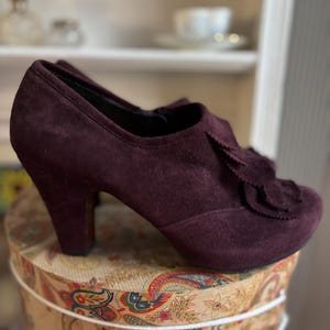 Op de afbeelding: Een paar bordeaux suède hakschoenen met een decoratief bloemdetail aan de zijkant.