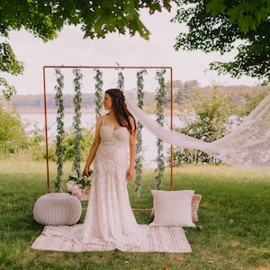 Könnte beinhalten: Eine Braut in einem weißen, bestickten Hochzeitskleid steht im Freien und hält einen Blumenstrauß. Ein durchsichtiger Schleier weht im Wind. Ein dekorativer Hintergrund mit Grün und einem Kupferrahmen befindet sich hinter ihr. Ein gewebter Teppich, ein Pouf und Kissen liegen auf dem Gras.