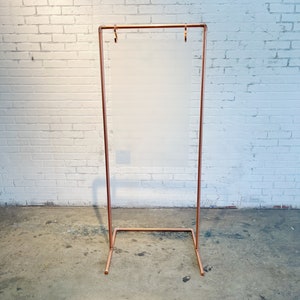 Puede incluir: Un soporte de letrero de metal de color cobre con un marco rectangular y un panel transparente esmerilado. El soporte del letrero tiene un diseño simple y minimalista y se encuentra sobre un piso de concreto contra una pared de ladrillo blanco.