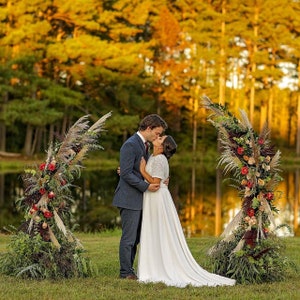 May include: A couple embraces and kisses under a floral archway during an outdoor wedding ceremony. The arch is decorated with pampas grass, red roses, and greenery. The background features a lake and trees with golden foliage.