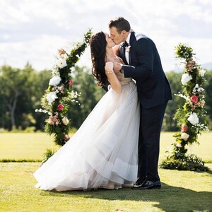 May include: A couple shares a kiss under a floral archway. The woman wears a white wedding dress with a layered skirt. The man is in a navy suit. The arch is decorated with white and pink flowers and greenery, set on a green lawn.