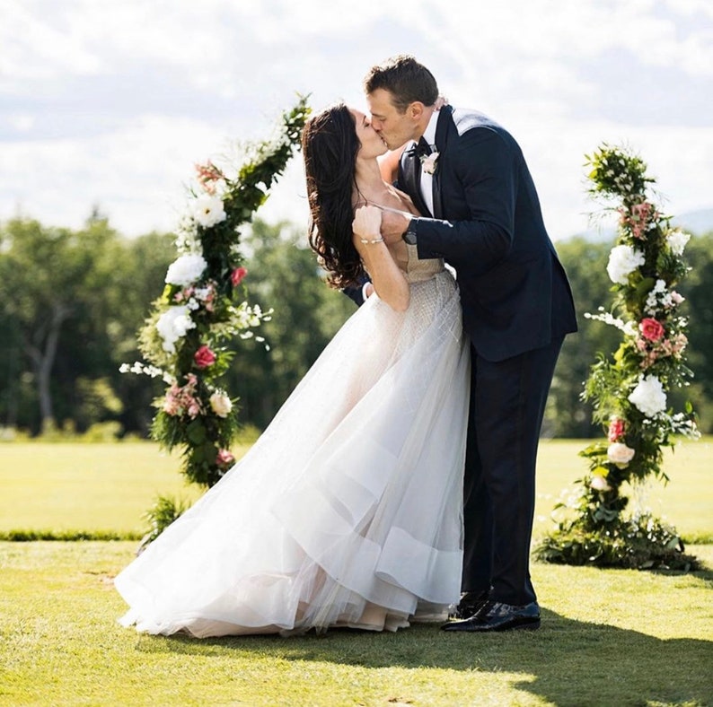 May include: A couple shares a kiss under a floral archway. The woman wears a white wedding dress with a layered skirt. The man is in a navy suit. The arch is decorated with white and pink flowers and greenery, set on a green lawn.