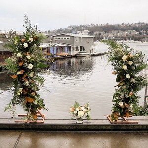 May include: An outdoor wedding arch with floral arrangements. Two copper-framed structures are adorned with white and peach flowers, green ferns, and foliage. A small bouquet sits in front of the arch, with a lake and houses in the background.