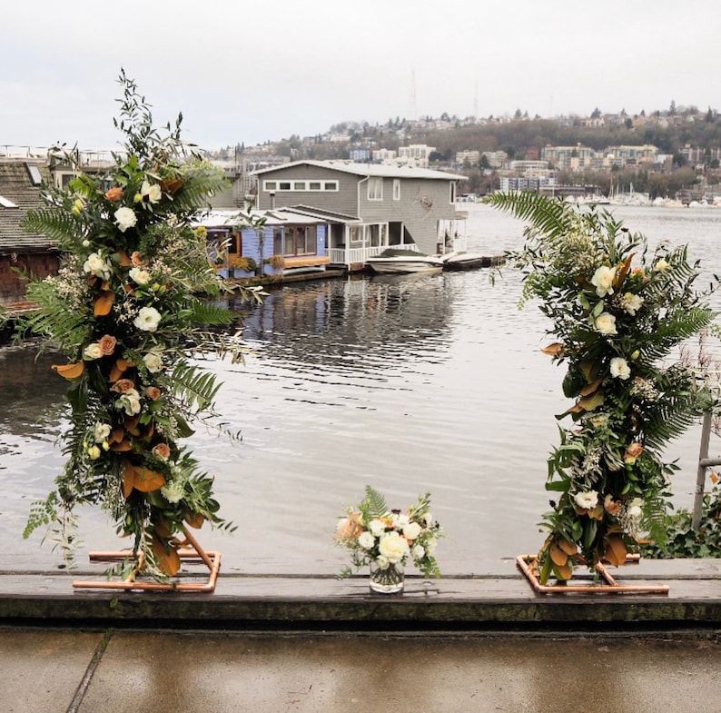May include: An outdoor wedding arch with floral arrangements. Two copper-framed structures are adorned with white and peach flowers, green ferns, and foliage. A small bouquet sits in front of the arch, with a lake and houses in the background.