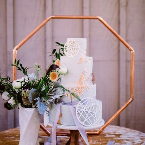 Könnte beinhalten: Ein dreistöckiger weißer Kuchen mit Goldblattakzenten und geometrischen Dekorationen steht auf einem Kuchenständer aus Holz. Ein geometrischer Kupferrahmen umgibt den Kuchen. Eine weiße Vase enthält ein Blumenarrangement mit Grün und Blumen. Hellblaue Bänder sind um den Kuchen drapiert.