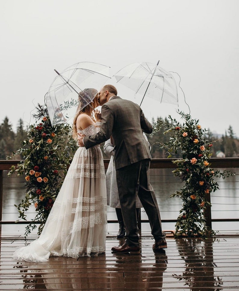 May include: A wedding ceremony takes place outdoors under clear umbrellas. The bride wears a white lace gown, and the groom wears a gray suit. Floral arrangements with orange and red flowers decorate the archway. The ceremony is taking place on a wooden deck.