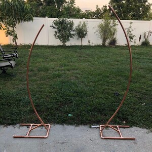 May include: Two copper-colored metal arch frames, each with a curved top and a rectangular base. The frames are set up on a concrete surface in a grassy backyard, suitable for weddings or events.