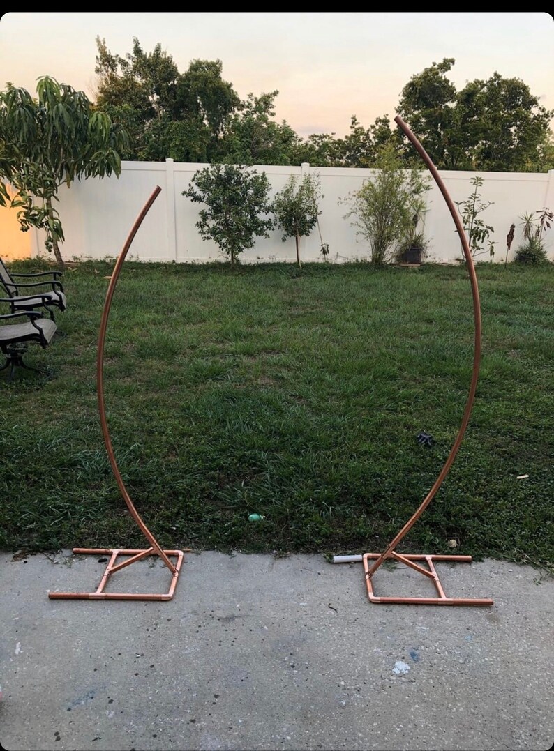 May include: Two copper-colored metal arch frames, each with a curved top and a rectangular base. The frames are set up on a concrete surface in a grassy backyard, suitable for weddings or events.