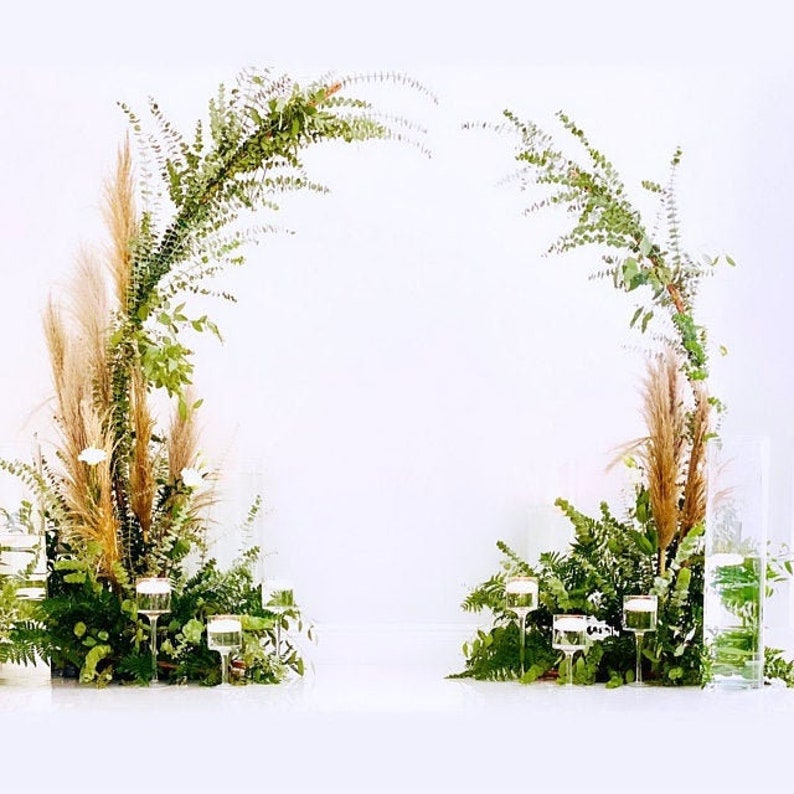 May include: An archway decorated with greenery and pampas grass, ideal for a wedding or event. The arch is adorned with lush green ferns and other foliage, with tall, feathery pampas grass plumes. Clear glass candle holders and vases add to the elegant decor.