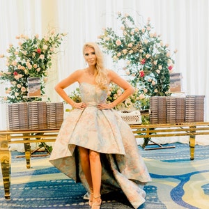 May include: A strapless, high-low formal gown with a floral pattern in pastel colors. The dress is worn by a person standing in front of a table with stacks of books and floral arrangements. The table is gold-colored.