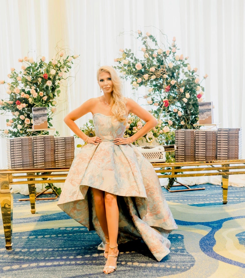 May include: A strapless, high-low formal gown with a floral pattern in pastel colors. The dress is worn by a person standing in front of a table with stacks of books and floral arrangements. The table is gold-colored.